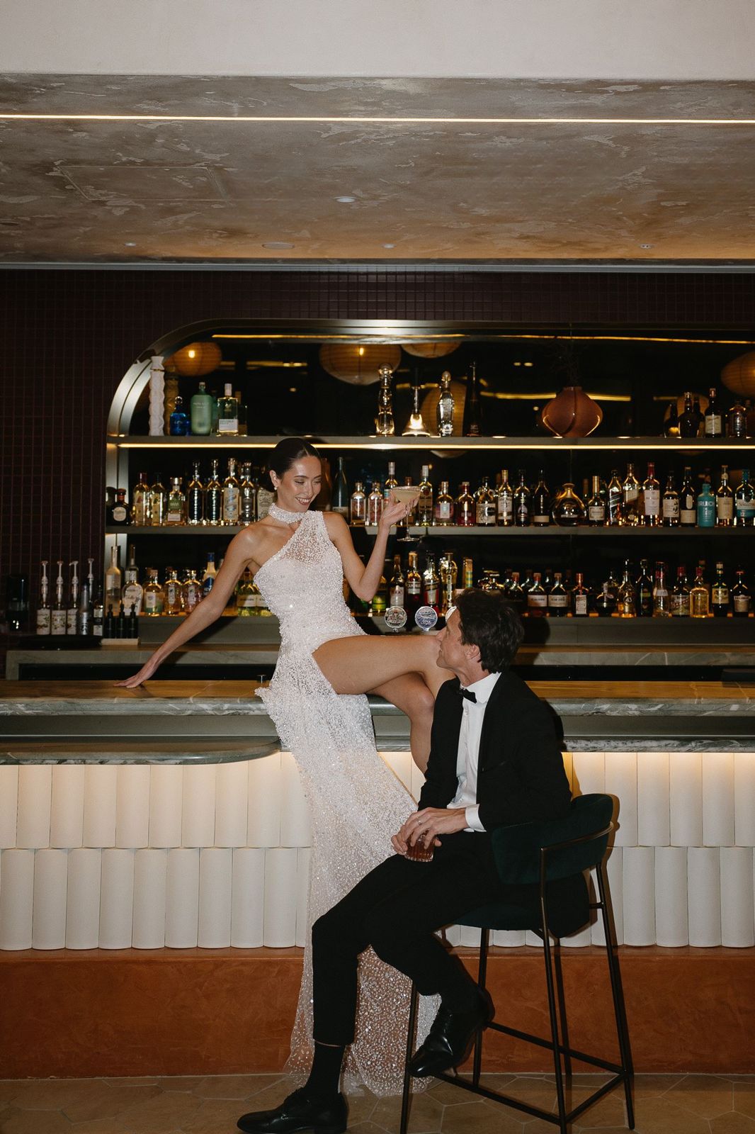 The bride perched on the bar with a cocktail as the groom looks up at her